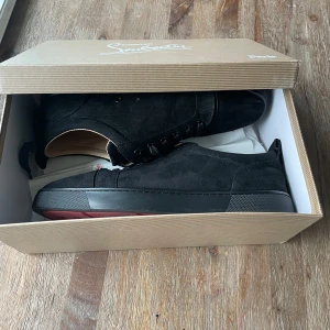 Christian Louboutin skor - Snygga svarta sneakers i mocka från Christian Louboutin med klassisk röd sula och svarta snören. Skorna har en låg profil och rund tå, perfekt för dig som vill ha en stilren men ändå lyxig look. Ikonisk signatur på sulan och diskreta detaljer. Box och kvitto m.m