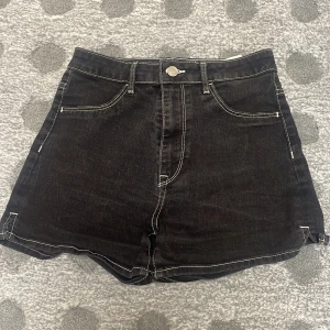 Svarta högmidjade jeansshorts - Svarta högmidjade jeansshorts med kontrasterande vita sömmar och klassisk fickdesign. Snygg knappstängning framtill och raka ben. Perfekta för en chill sommarlook eller när du vill ha en enkel men stilren vibe.