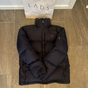 Svart dunjacka Polo Ralph Lauren - Säjer denna Snygg svart dunjacka från Polo Ralph Lauren med klassisk röd logga på bröstet pågrund av att jag vuxit ur den. Jackan har hög krage, dragkedja framtill och flera fickor med dragkedja. Fylld med 80% dun och 20% fjäder, yttertyg i polyester. Perfekt för kalla dagar och riktigt stilren look. 