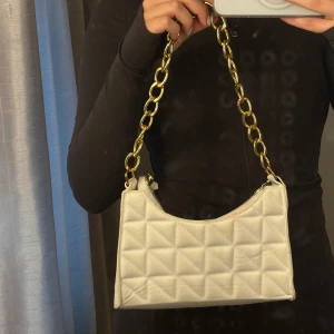Vit quiltad axelväska med guldkedja - Säljer en snygg vit axelväska med quiltat mönster och en chunky guldfärgad kedja som axelrem. Väskan har dragkedja upptill och en stilren, modern look som passar till många outfits.