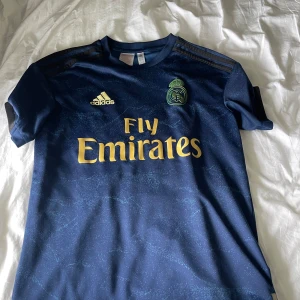 Real Madrid adidas marinblå tröja L - Snygg marinblå Real Madrid fotbollströja från adidas med guldiga detaljer och Fly Emirates-tryck. Tröjan har korta ärmar, klubbmärke på bröstet och diskret mönster i tyget. Perfekt för dig som älskar fotboll och vill sticka ut på planen.