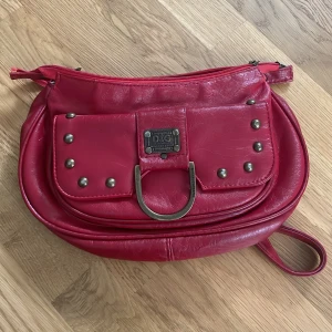 Röd väska från Dolce & Gabbana - Röd Väska från Dolce & Gabbana i skinn med metall-detaljer. Väskan har en dragkedja upptill och en framficka med D&G-plakett. Perfekt storlek för det viktigaste och en riktigt statement piece. Dragkedjan är lite defekt i vänstra hörnet men annars är den hel. Köpt secondhand så oklart om den är äkta vill inte chansa så säljer den billigt🩷