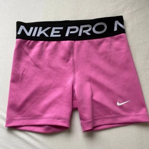 Nike Pro rosa träningstights shorts - Original pris: 239kr. Snygga rosa Nike Pro shorts i tight passform med svart resår i midjan och stor logga. Tillverkade i Dri-FIT material som andas och håller dig torr under träningen. Perfekta för gymmet eller löprundan, med klassisk vit swoosh på benet.