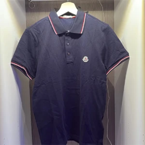Mörkblå Moncler pikétröja - Pris kan diskuteras!!!Snygg mörkblå piké från Moncler med klassisk krage och tre knappar framtill. Tröjan har korta ärmar och detaljer i rött, vitt och blått på krage och ärmslut. Ikonisk Moncler-logga broderad på bröstet. Perfekt för dig som gillar stilrena och exklusiva plagg.