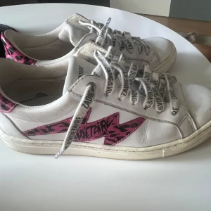 Zadig & Voltaire vita sneakers med rosa detaljer - Snygga vita sneakers från Zadig & Voltaire med  rosa och svarta blixtdetaljer på sidorna och hälen. Små defekter här o var men allt syns på bilderna! Pris kan diskuteras 