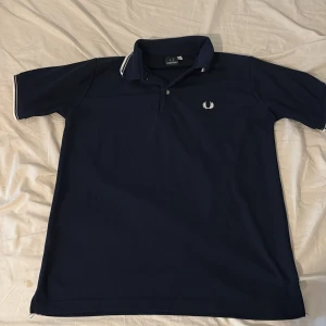 Mörkblå Fred Perry pikétröja L - Snygg mörkblå pikétröja från Fred Perry i storlek L. Klassisk design med vit logga broderad på bröstet och vita detaljer på krage och ärmslut. Tröjan har korta ärmar och är gjord i mjuk bomull, perfekt för en clean och sportig look.