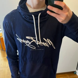Mörkblå hoodie i merino - Säljer en snygg mörkblå hoodie från Mons Royale med vitt tryck på framsidan. Tröjan har en justerbar huva med vita snören och en stor ficka framtill. Perfekt för en avslappnad stil. Tröjan är i 91% merino. Skicket är utmärkt och inge defekter eller liknande förekommer.