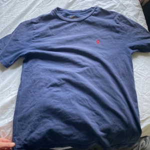 Blå t-shirt från Polo Ralph Lauren - Säljer en klassisk blå t-shirt från Polo Ralph Lauren med det ikoniska röda broderade logotypen på bröstet. Perfekt för en stilren och avslappnad look. Tillverkad i mjukt material för extra komfort.