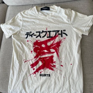 Dsquared2 T-shirt - Säljer en cool vit t-shirt från Dsquared2 med ett iögonfallande rött tryck och japansk text på framsidan. Perfekt för dig som vill sticka ut med en unik stil. T-shirten har korta ärmar och är gjord i mjukt bomullsmaterial. Lite skavanker som syns på bilder.