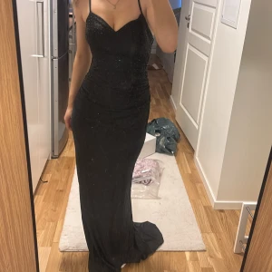 Svart glittrig balklänning - Elegant svart långklänning med smala axelband och glittrande detaljer. Klänningen har en figurnära passform och en v-ringad urringning. Perfekt för bal. Köptes för 1900kr på Zapaka 