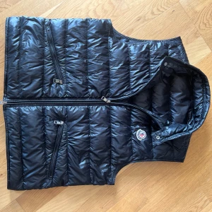 Moncler Gui väst - Snygg svart Moncler Gui väst med dragkedja och flera praktiska fickor. Perfekt för lager-på-lager under kyliga dagar. Västen har en quiltad design och en stilren look. Storlek 2 vilket motsvarar en liten M ungefär. Inga större defekter förutom att den har använts. Vid köp följer endast västen med då jag tappat bort kvitto och box. Hör av er vid frågor!🙌🏻