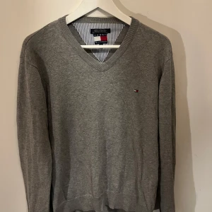 Grå v-ringad tröja från Tommy Hilfiger - Säljer en stilren grå v-ringad tröja från Tommy Hilfiger. Tröjan har långa ärmar och en liten logotyp på bröstet. Perfekt för en avslappnad men snygg look. Skriv för fler bilder! 