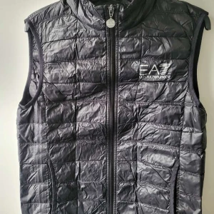 EA7 väst  - Snygg svart quiltad väst från Emporio Armani med dragkedja framtill. Perfekt för lager-på-lager och stilren look. EA7-logga på bröstet ger en exklusiv touch.