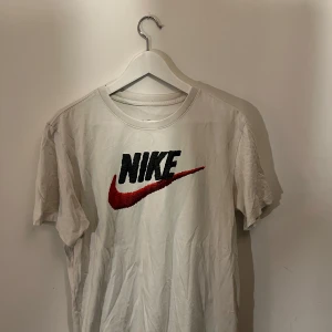 Vit kortärmad t-shirt från Nike - Säljer en klassisk vit t-shirt från Nike med det ikoniska logotypen i svart och rött på framsidan. Perfekt för en sportig look eller avslappnad stil. T-shirten är kortärmad och gjord i bomull för en bekväm passform.