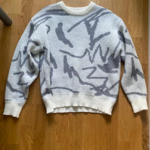 Vit och grå stickad tröja - Säljer en vit stickad tröja med grått abstrakt mönster. Tröjan har långa ärmar och ribbade muddar vid ärmslut. Skriv om du har frågor!