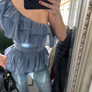 Blå oneshoulder blus med volanger - Säljer en snygg blå oneshoulder blus med volanger. Perfekt för en stilren look. Blusen har en elastisk midja som ger en fin passform och är tillverkad i ett lätt och luftigt material.
