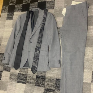 Grå kostym från Riley - Snygg grå kostym från Riley med kavaj och byxor. Kavajen har två knappar och klassisk passform. Två slipsar ingår, en svart och en med paisleymönster. Perfekt för formella tillfällen.
