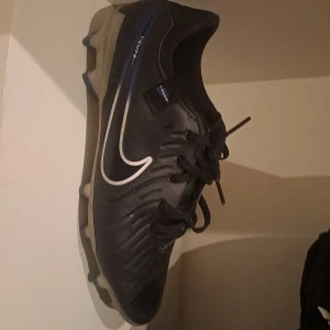 Svarta fotbollsskor från Nike storlek 39 - Säljer ett par svarta Nike fotbollsskor med vit swoosh och snörning. Skorna har en rund tå och är designade för optimal prestanda på planen. Perfekta för fotbollsträning och matcher.