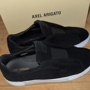 Svarta sneakers från Axel Arigato - Snygga svarta sneakers från Axel Arigato i mocka med elastiska band över vristen för enkel påtagning. Skorna har en vit sula som ger en stilren kontrast. Perfekta för en avslappnad men trendig look. Köptes i februari men har knappt används, de är även sällsynta eftersom dem inte säljs mer. Byten funkar. Mvh Oliwer
