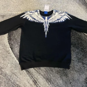  Marcelo Burlon - Snygg svart tröja från Marcelo Burlon med ett iögonfallande vitt och blått vingmönster över axlarna. Perfekt för dig som vill sticka ut med en unik design. Tröjan har långa ärmar och en bekväm passform.