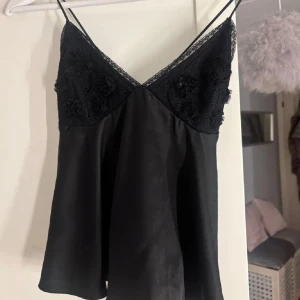 Svart linne med spetsdetaljer - Elegant svart linne med tunna axelband och vacker spetsdetalj vid halsringningen. Toppen har en lös passform och knytning i ryggen för extra stil. Perfekt för en chic och sofistikerad look. Ifrån Zara i storlek S, fler bilder finns ej då den ej passar mig