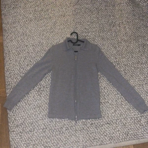 Grå tröja med dragkedja från J.Lindeberg - Säljer en stilren grå tröja från J.Lindeberg med dragkedja framtill. Tröjan har en klassisk krage och långa ärmar, perfekt för en avslappnad men snygg look. Passar bra till både jeans och chinos.