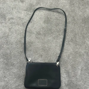 Svart axelväska med dragkedja - Snygg svart axelväska i skinn med justerbar rem och dubbla dragkedjor. Perfekt för att hålla dina saker säkra och organiserade. Stilren design med en diskret logotyp på framsidan. Är från Marc Jacobs och köpt för flera är sedan. Ordpris var ca 3000kr men säljs för 800kr. Pris går att diskuteras!