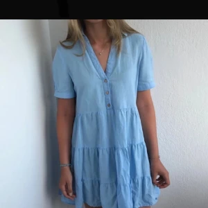 Ljusblå klänning med knappar - Charmig ljusblå klänning med korta ärmar och v-ringning. Klänningen har knappar framtill och volanger som ger en luftig känsla. Perfekt för varma dagar!