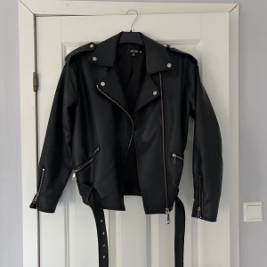 Svart skinnjacka med dragkedja - Snygg svart skinnjacka med dragkedja och bälte. Jackan har långa ärmar och flera silverfärgade detaljer som knappar och dragkedjor. 