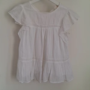 Vit blus med spetsdetaljer - Charmig vit blus med korta ärmar och vackra spetsdetaljer. Blusen har en luftig och lätt design med volanger som ger en romantisk touch. Perfekt för varma sommardagar.