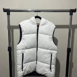 Vit dunväst från Guess - Snygg vit dunväst från Guess med huva och dragkedja. Västen har svarta detaljer och två fickor med dragkedja framtill. Perfekt för lager-på-lager under kyligare dagar. En söm i högra fickan har öppnat sig men påverkar inte funktionen och inget ramlar ut ur fickan