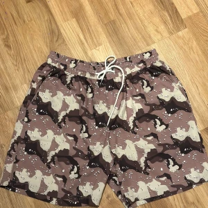 Camouflage shorts med vit snörning - Camouflage mönster med jordnära färger. De har en bekväm passform med elastisk midja och snörning för justerbarhet. Köpt 2024 på en marknad. 