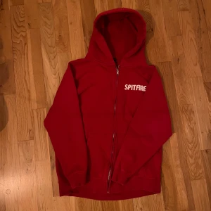 Röd hoodie från Spitfire - Säljer en cool röd hoodie från Spitfire med dragkedja och huva. Framsidan har en vit logotyp och baksidan pryds av ett stort, tufft tryck. Perfekt för skateparken eller chill dagar.