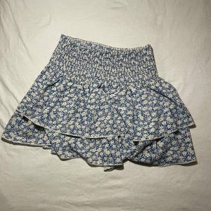 Blommig kjol med volang - Säljer en söt kjol i blått och vitt med blommigt mönster. Kjolen har en volangdetalj och en elastisk midja för en bekväm passform. Pris kan diskuteras!