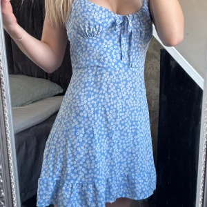 Blå blommig klänning - Söt blå klänning med vita blommor och volangdetalj längst ner. Klänningen har en knytning framtill och är ärmlös. Dragkedja bak i ryggen och knytningar vid axlarna som man kan knyta själv. Står en någon storlek men sitter som xs/xxs. 