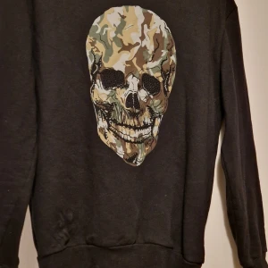 Svart tröja med dödskalle från Zara (oanvänd) - Säljer en svart tröja från Zara med ett stort dödskalletryck i camouflage på framsidan. Tröjan har långa ärmar och är perfekt för en cool och avslappnad stil. Passar bra till jeans eller cargo byxor.Oanvänd