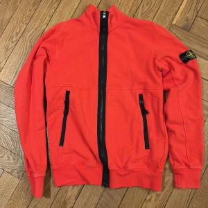 Röd jacka från Stone Island - Snygg röd jacka från Stone Island med svarta dragkedjor och en ikonisk logga på ärmen. Jackan har långa ärmar och praktiska fickor med dragkedja. Perfekt för en stilren look.