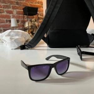 Svarta solglasögon från Ray-Ban - Snygga svarta solglasögon från Ray-Ban med klassisk design. De har en rektangulär form med lila glas som ger ett stilrent intryck. Använda utomlands i somras, små repar på glaset, och små scratches på bågarna men annars väldigt trevliga.