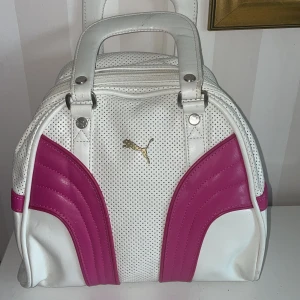 Vit och rosa handväska från Puma - Snygg vit handväska från Puma med rosa detaljer och perforerat mönster. Små läder bortfall.Väskan har ett guldigt Puma-märke och dragkedja med logotyp. Perfekt för att ge en sportig touch till din stil.