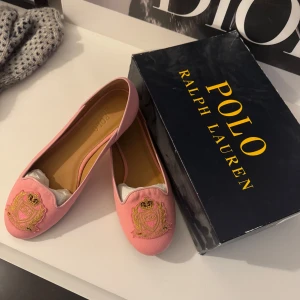 Rosa loafers från Polo Ralph Lauren - Säljer ett par stiliga rosa loafers från Polo Ralph Lauren. Skorna har en broderad detalj framtill med ett elegant emblem i guld och rosa. Perfekta för en chic och avslappnad look.