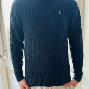 Mörkblå stickad tröja från Ralph Lauren - Säljer en snygg mörkblå stickad tröja från Ralph Lauren med kabelstickat mönster och det klassiska logotypbroderiet i orange på bröstet. Tröjan har långa ärmar och ribbade kanter för en stilren look. Perfekt för kyligare dagar!