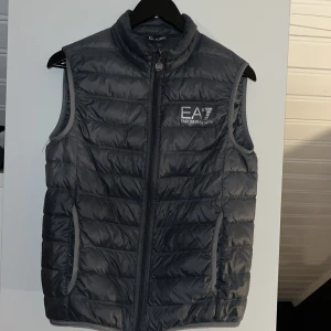 Grå dunväst från Emporio Armani - Snygg grå stilren dunväst från Emporio Armani med dragkedja och hög krage i ett mycket gott skick. Finns två ytter fickor och två innerfickor på denna dunväst. Tags, påse till västen följer också med. Vänligen hör av er vid frågor, svarar snabbt!!!😁