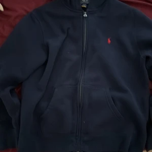 Mörkblå hoodie från Polo Ralph Lauren - Säljer en snygg mörkblå hoodie från Polo Ralph Lauren med dragkedja och det klassiska röda logotypen på bröstet. Perfekt för en avslappnad stil. Den har långa ärmar och en bekväm huva. Passform 160-166