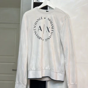 Vit långärmad tröja från Armani Exchange - Säljer en stilren vit långärmad tröja från Armani Exchange med deras logotyp tryckt på framsidan. Tröjan har en rund halsringning och ribbade muddar vid ärmslut och nederkant. Perfekt för en avslappnad och trendig look.