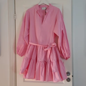 Rosa klänning från Neo Noir - Säljer en söt rosa klänning från Neo Noir med volanger och knytband i midjan. Klänningen har långa ärmar och en v-ringad hals. Perfekt för en somrig look!