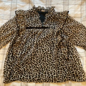 Leopardmönstrad blus med volanger från LINDEX!  - Snygg leopardmönstrad blus med volanger och knappar framtill. Blusen har långa ärmar och en v-ringad design. Perfekt för att ge en trendig touch till din outfit. Blusen är använd ganska mycket men har nästan inga täcken på användning! Den är i storlek 158 och skriv för fler frågor! 💕 