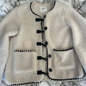 Vit teddyjacka med svarta detaljer   - Mjuk vit teddyjacka med svarta detaljer. Jackan har knappar och dekorativa snörningar framtill samt två praktiska fickor. Perfekt för kyliga dagar! Säljer vidare  eftersom jag köpte fel storlek, har en svart fläck på armen som jag inte visste om och har ej testat att ta bort men annars väldigt fin och ren! 