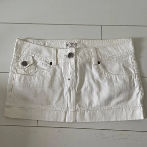 Vit kortkjol med fickor - Snygg vit kortkjol med klassisk jeansdesign. Kjolen har fram- och bakfickor med dragkedjor och en dekorativ bältesdetalj baktill. Perfekt för en stilren look.