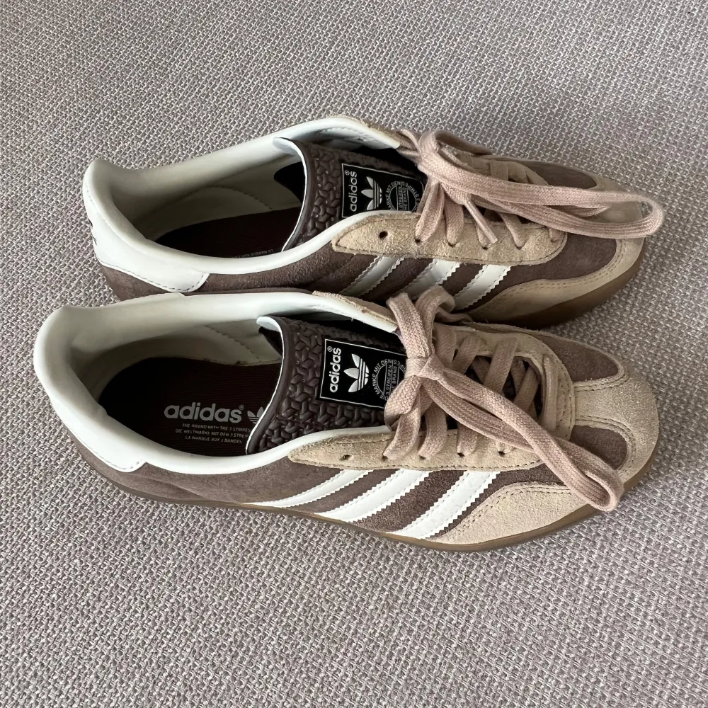 Säljer ett par klassiska Adidas Gazelle sneakers i beige och brunt. Skorna är tillverkade i mocka med vita skinn detaljer och har den ikoniska tre-randiga designen. Perfekta för en stilren look. . Kengät.
