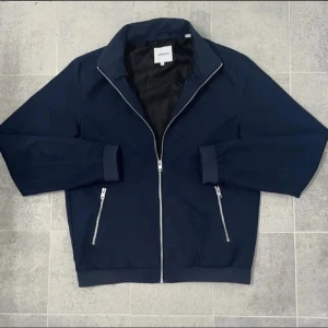 Mörkblå jacka med dragkedja - Snygg mörkblå jacka från jack n jones med dragkedja framtill och två fickor med dragkedjor. Jackan har en stilren design med långa ärmar och ribbade muddar vid ärmslut och nederkant.